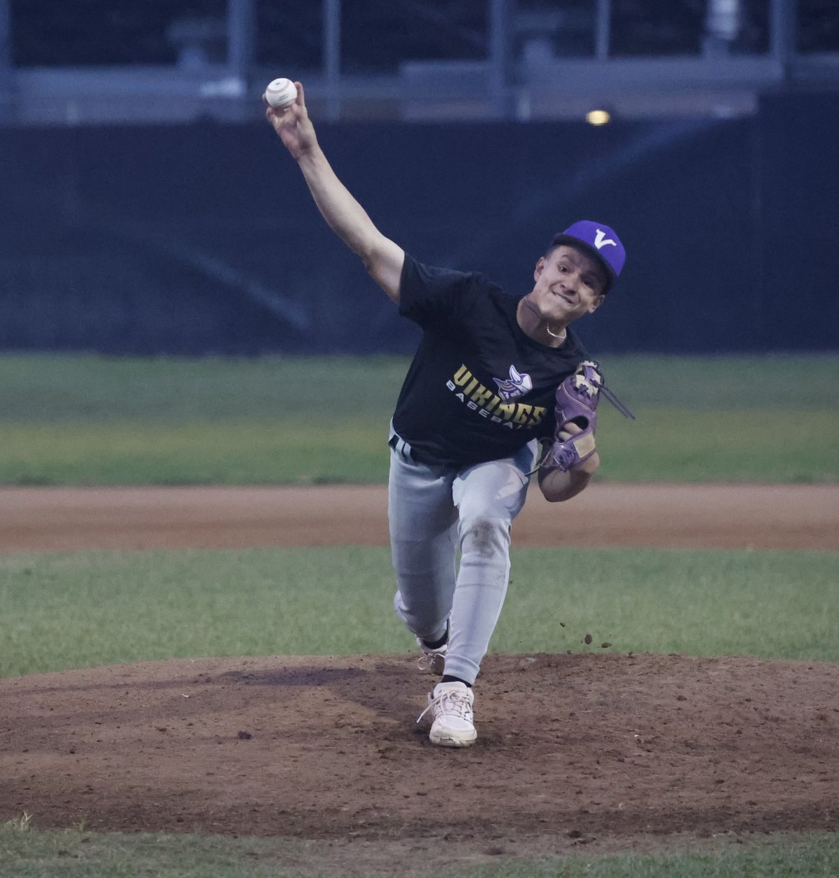 Miles Shelofsky at the top of his delivery on the mound under the lights, the ball still in his right hand at release, his Vikings cap and jersey dark against the outfield wall behind him.