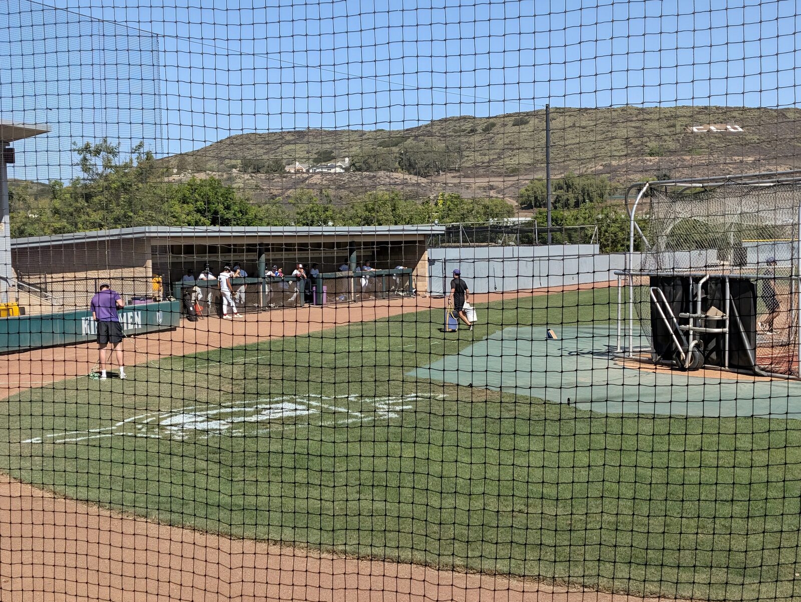 A covered dugout during batting practice, with the CLU letters on the hillside behind the field.