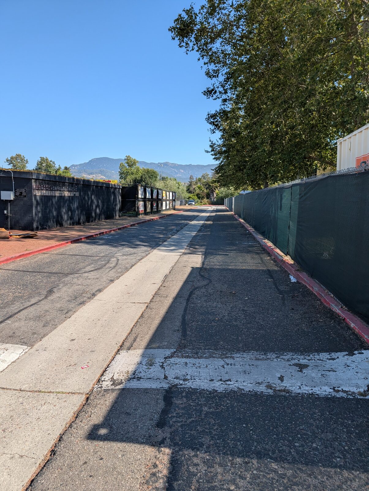 The access path behind the third-base dugout, between the dumpsters and the field perimeter, hills beyond the outfield.